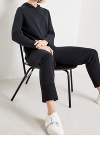 Schwarzer Kapuzenpullover mit Kordelzug und passender tapered Hose, kombiniert mit weißen Plateauschuhen. Der Stoff wirkt glatt mit gerippten Bündchen.