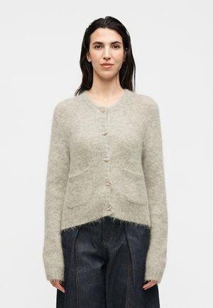 Femme aux cheveux foncés portant un cardigan gris clair duveteux boutonné et un pantalon large taille haute foncé, debout devant un fond uni.
