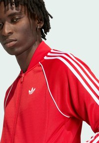 Mladý človek s tmavými vrkôčkami, ktorý má na sebe červenú športovú bundu na zips s bielymi pruhmi a logom na ramene a hrudi.