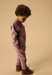 Bambino con capelli ricci che indossa un pullover e pantaloni color malva, che guarda in basso, in piedi contro uno sfondo beige.