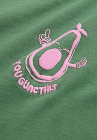 Zöld pamut anyag, pink hímzett avokádóval, amelynek karjai vannak, kezét a levegőbe emelve, alatta a "TE GUAC TETTÉL" felirattal játékos betűtípussal.