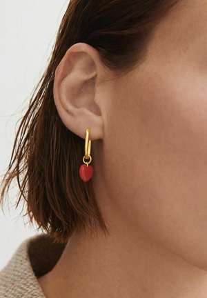 Zelta stīpiņas auskars ar mazu sarkanu sirsniņas piekariņu, kas valkāts kreisajā ausī persona ar īsiem brūniem matiem un gaišu ādu.