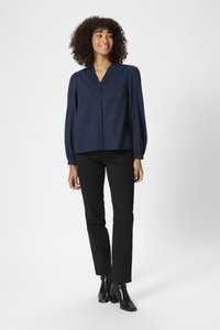 Blouse marine à boutons sur le devant, décolleté en V et encolure texturée ; associée à un jean noir à jambes droites et des bottines à talons noires.