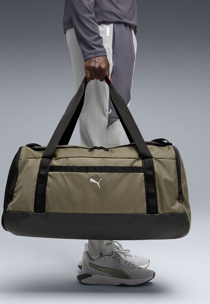 Sac polochon vert olive avec accents et poignées noirs, doté d'une poche à fermeture éclair à l'avant et d'un logo blanc. Texture lisse, forme rectangulaire.