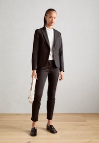 Černý upravený blazer přes bílé tričko, doplněný černými slim-fit kalhotami a lesklými polobotkami. Model drží bílou tašku. Jednoduché pozadí.