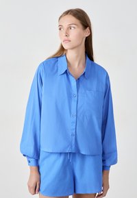 Jasnoniebieska koszula zapinana na guziki z kołnierzykiem, pojedynczą kieszenią na piersi i długimi bufkami, noszona z pasującymi szortami. Gładka tekstura, casualowy krój.