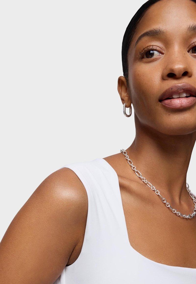 Top bianco senza maniche con scollatura a quadrato, abbinato a una collana di catena argento spessa e piccoli orecchini a cerchio argento. Texture della pelle liscia.