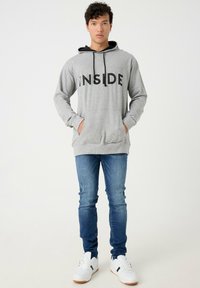 Sudadera gris con texto negro "INSIDE", bolsillo canguro y capucha con cordón, combinada con jeans azules y zapatillas blancas.