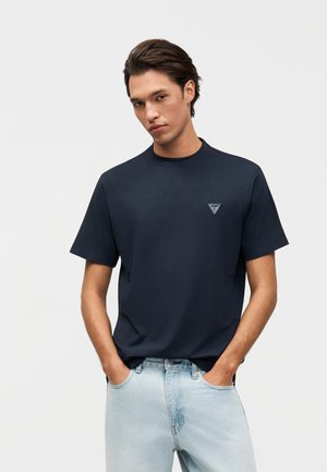 Jeune homme portant un t-shirt à manches courtes bleu marine avec un petit logo triangulaire sur la poitrine et un jean bleu clair, les mains dans les poches.