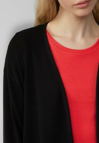Cardigan noir avec un design en V superposé sur un haut rouge côtelé. Le cardigan possède des manches longues et une texture douce.
