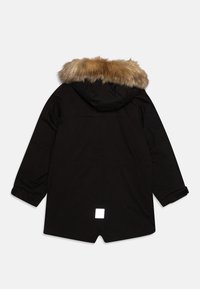 Parka nera con cappuccio foderato in pelliccia, polsini elasticizzati e una patch quadrata riflettente nella parte inferiore della schiena. Il tessuto ha una consistenza liscia.