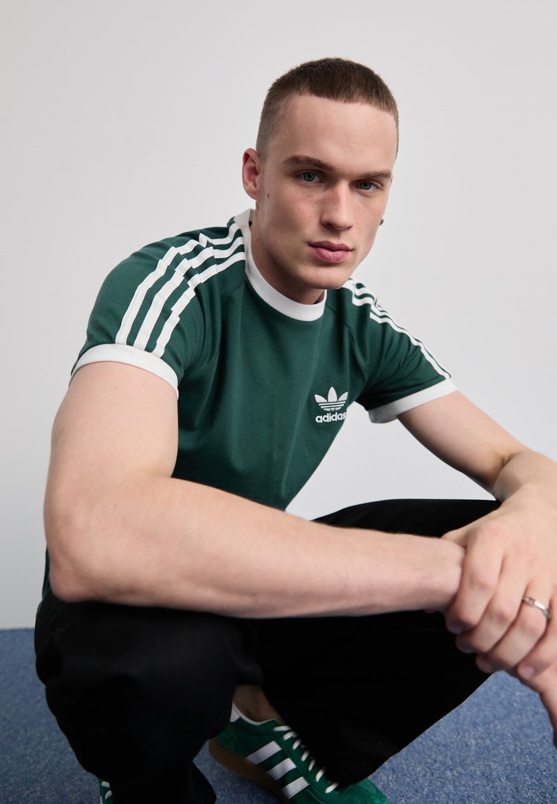 Žalias medvilnės marškinėlis su baltais kraštais ir trimis baltomis juostelėmis ant rankovių. Su "Adidas" logotipu. Modelis dėvi juodas kelnes ir žalias sportbačius.