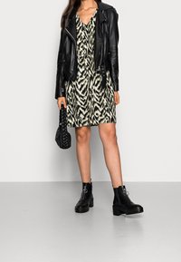 Black leather biker jacket over a patterned, knee-length dress in green and black. Matched with black ankle boots and a braided handbag.