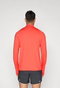 Αθλητική μπλούζα με μακριά μανίκια σε φωτεινό κόκκινο, κατασκευασμένη από ελαφρύ υλικό, με ψηλό γιακά και λευκές λεπτομέρειες στην πλάτη.