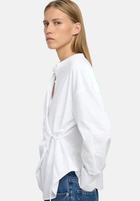 Biała, długosleeve koszula z wiązaniem, z wysokim dekoltem, szczegółem wiązania z tyłu oraz miękką fakturą. Połączona z niebieskimi dżinsami.