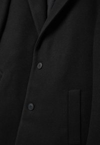 Abrigo de lana negra con tela texturizada, dos botones negros, un bolsillo en el pecho y un diseño de cuello con solapa.