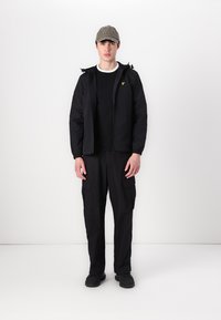 Schwarze Zip-Jacke mit elastischen Bündchen, kombiniert mit einem schwarzen T-Shirt und Cargo-Hosen. Eine graue Kappe mit dem Wort "Won" wird getragen.