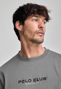 Sudadera gris con cuello redondo acanalado, que presenta el texto negro "POLO CLUB" y "ESTABLECIDO 2012" en dorado debajo. Textura suave, corte relajado.