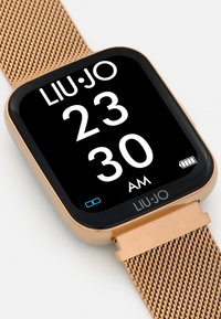 Smartwatch color oro con display quadrato nero, grandi numeri bianchi, cinturino in metallo a rete e un'icona della batteria nell'angolo in basso a destra.