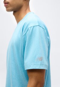 Πλάγια όψη ενός ατόμου που φοράει ανοιχτό μπλε αθλητική μπλούζα με κοντά μανίκια και ένα μικρό λογότυπο New Balance στο μανίκι.