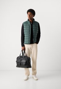 Grüne, flauschige Weste, schwarzer Pullover, cremfarbene Hose und weiße Sneaker. Hält eine schwarze Ledertasche. Glatte Textur und minimalistisches Design.