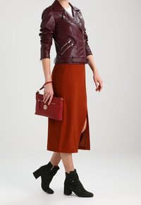 Kobieta w bordowej skórzanej kurtce, miedzianej midi spódnicy z rozcięciem, czarnych botkach za kostkę, trzymająca pasującą bordową torebkę kopertową.