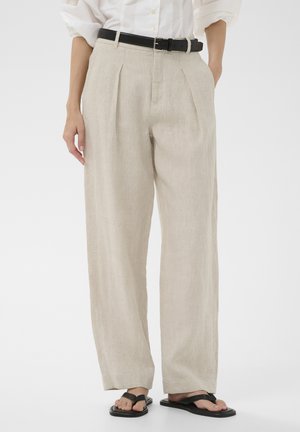 Femme portant un pantalon ample en lin beige avec des plis sur le devant, une ceinture noire, une chemise blanche et des sandales plates noires, debout sur un fond blanc.