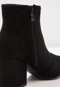 Bottine noire en matériau semblable au daim, dotée d'une fermeture éclair latérale, d'un talon bloc, et d'une texture lisse avec des détails de coutures.