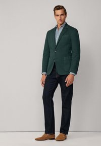 Blazer en coton vert avec des revers à cran, deux poches avant et une coupe slim, associé à une chemise rayée, un jean en denim foncé et des mocassins beige.