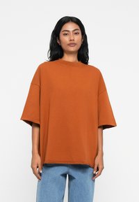 Oversized μπλούζα χρώματος σκουριάς από μαλακό ύφασμα με κοντά μανίκια, στρογγυλή λαιμόκοψη και ορατές ραφές. Συνδυασμένη με ανοιχτές μπλε τζιν.