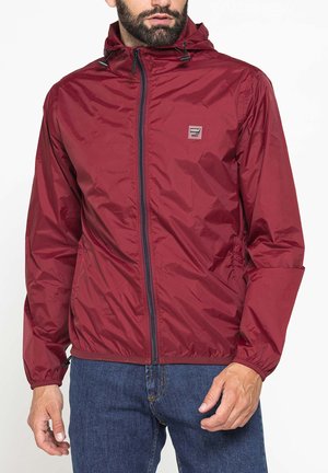 Uomo che indossa una giacca a vento con cappuccio color granata, con zip frontale e polsini elastici, abbinata a jeans blu.