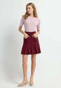 Lichtroze top met korte mouwen gecombineerd met een bordeauxrode geplooide rok met twee voorzakken. Het model draagt beige lage hakken met accenten.
