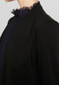 Mustapuku, jossa on teksturoitu laivastonsininen pitsikaulus, jossa on risat reunat. Muotoilussa on räätälöidyt olkapäät ja siisti siluetti.