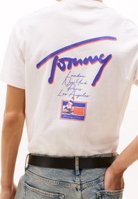Biały bawełniany t-shirt z dużym niebiesko-czerwonym logo "Tommy" na plecach, z nazwami miast i grafiką żaglówki na środku.