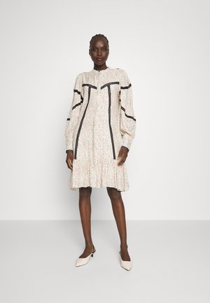Robe à motif floral avec de longues manches bouffantes, des accents noirs, un ourlet volants et un col à boutons. Portée avec des chaussures à talons de couleur claire.