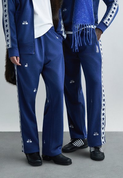 Pantalon de survêtement bleu avec imprimés de lapins blancs et étoiles sur les côtés, fabriqué en tissu doux avec une coupe décontractée, associé à des chaussures noires.
