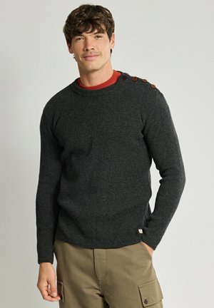 Uomo che indossa un maglione grigio scuro con bottoni sulle spalle sopra una camicia rossa e pantaloni cargo beige, in piedi con una mano in tasca.