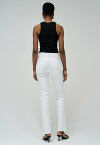 L'image montre une personne portant un débardeur noir ajusté et un jean blanc moulant. Le jean dispose de poches arrière et d'un design minimaliste.