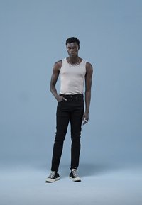 Pánské béžové tílko, černé džíny s úzkým střihem a černé tenisky. Model stojí před světle modrým pozadím a nosí stříbrný řetízek.