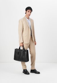 Schwarze Leder-Tote mit doppelten Griffen, vertikalen Nähten und einem abnehmbaren Anhänger. Vor einem neutralen Hintergrund mit einer im Anzug gekleideten Person.