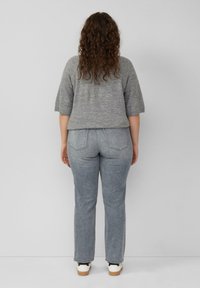 Osoba z kręconymi brązowymi włosami, ubrana w szary sweter z krótkim rękawem, wyblakłe niebieskie dżinsy i białe trampki, stojąca na jednolitym tle.