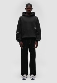 Czarna, oversize bluza z kapturem z przednią kieszenią, ozdobiona białymi haftowanymi detalami. W połączeniu z czarnymi spodniami z szerokimi nogawkami oraz kolorowymi trampkami.