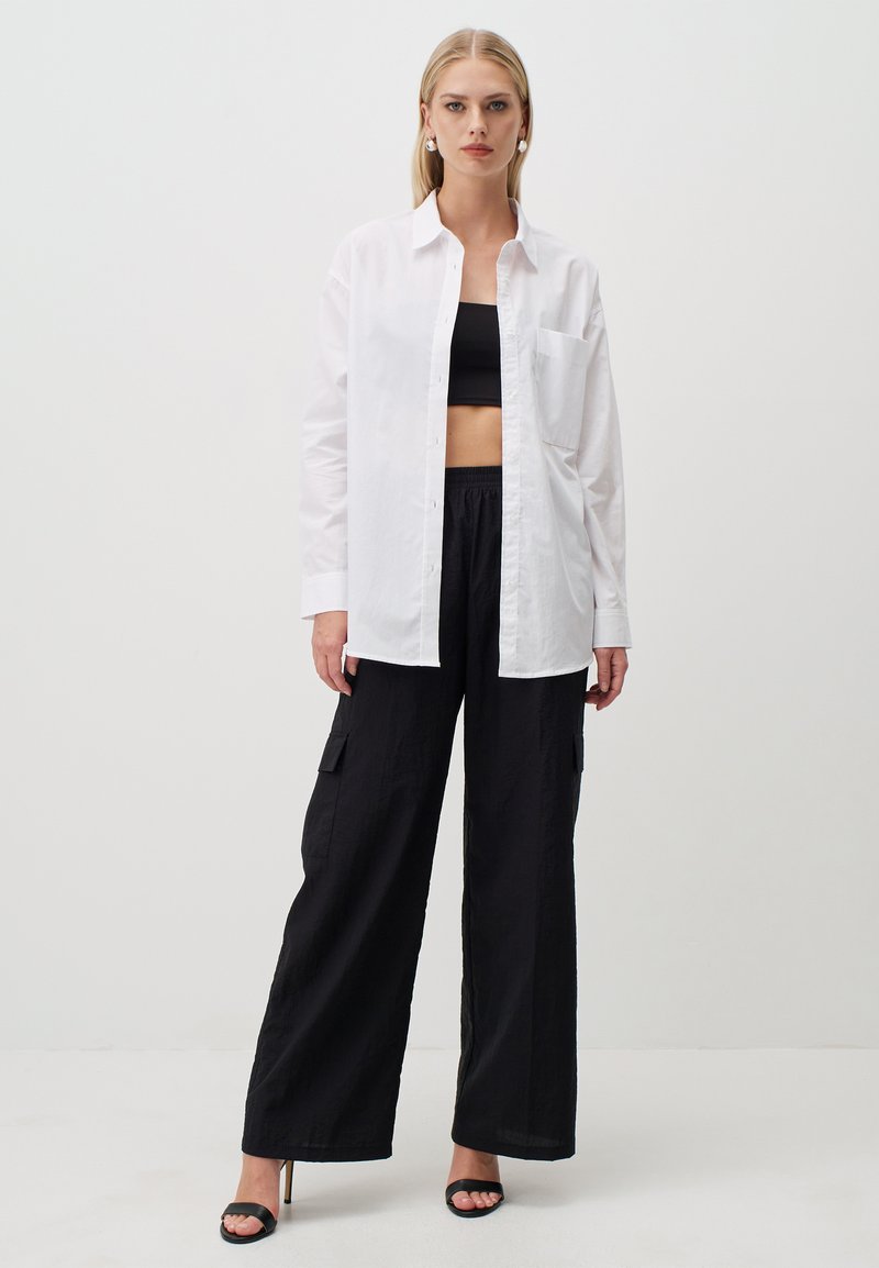 Witte button-up shirt over een zwart crop top, gecombineerd met wijde zwarte cargobroek. Het shirt heeft een borstzak en opgerolde mouwen.