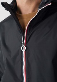 Zwarte windjack met een voorrits voorzien van rood-wit-blauwe accenten en een ronde metalen trekker. Glad, lichtgewicht stof.