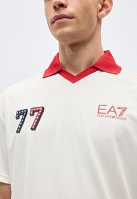 Άνδρας που φοράει λευκό πουκάμισο με κόκκινο γιακά, με κεντητό σήμα με τον αριθμό 77 και αστέρια στο αριστερό στήθος και κόκκινο λογότυπο EA7 Emporio Armani στο δεξί στήθος.