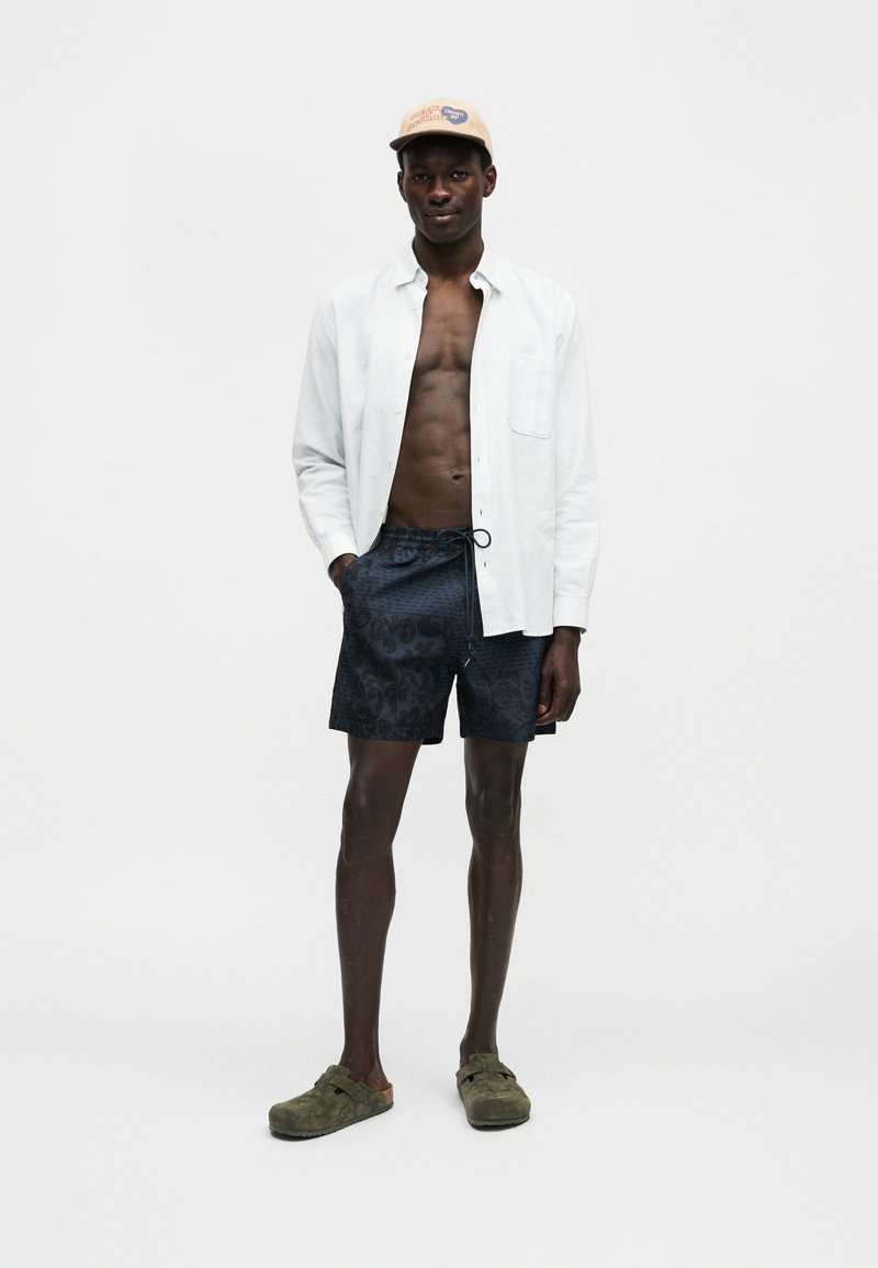 Mann trägt eine beige Mütze, offenes weißes Hemd, gemusterte dunkle Shorts und olivgrüne Slipper und steht mit einer Hand in der Hosentasche.