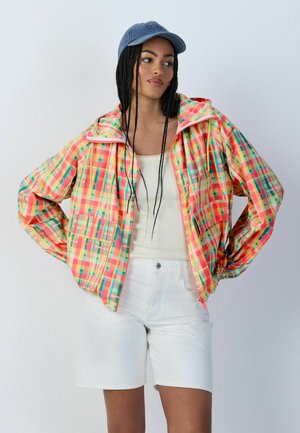 Naine punutud juustega, kes kannab sinist nokamütsi, erksavärvilist ruudulist jopet, valget topi ja valgeid lühkse, seisab käed puusadel.