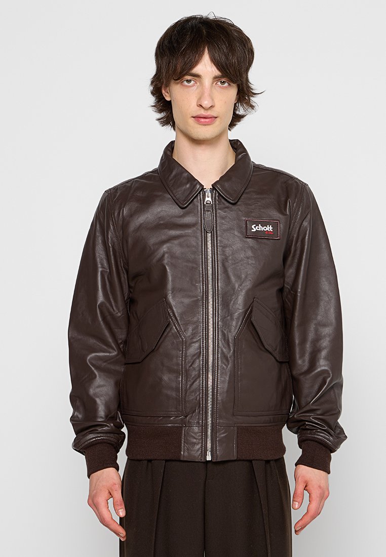 Veste bombardier en cuir marron avec fermeture éclair, deux poches avant et poignets côtelés. Présente un patch avec logo sur la poitrine gauche.