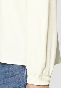 Blouse blanche texturée à manches longues et poignets élastiques, présentant une coupe lâche et un ourlet légèrement courbé, associée à un jean en denim bleu.