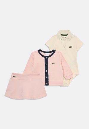 Tri oblačila za otroke: roza rebrasta cardigan z mornarskimi gumbi, kremna polo majica in roza krilo z detajlom logotipa.